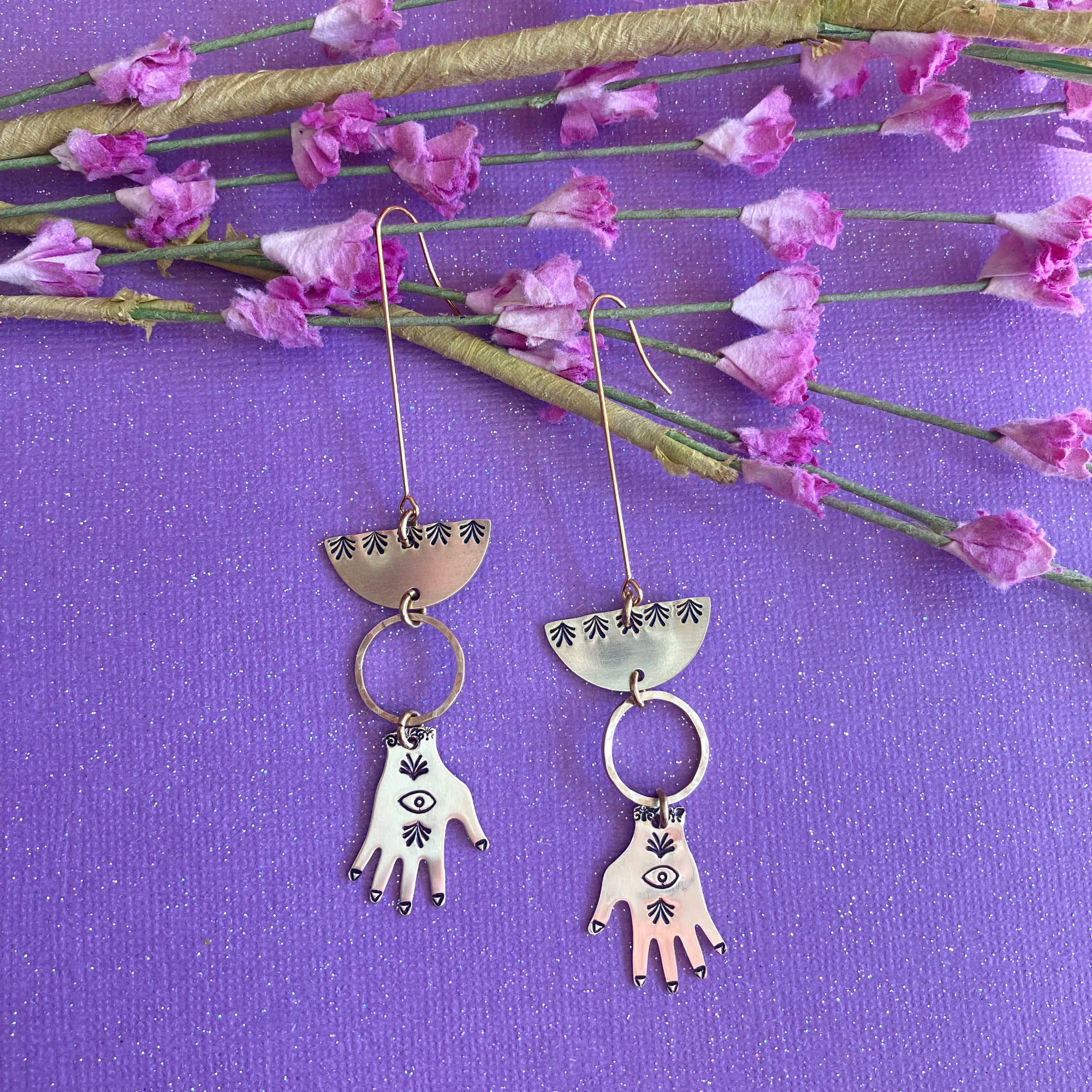 A pair of handmade brass hand earrings with engraved metal charms, displayed on a textured purple background with pink flowers.