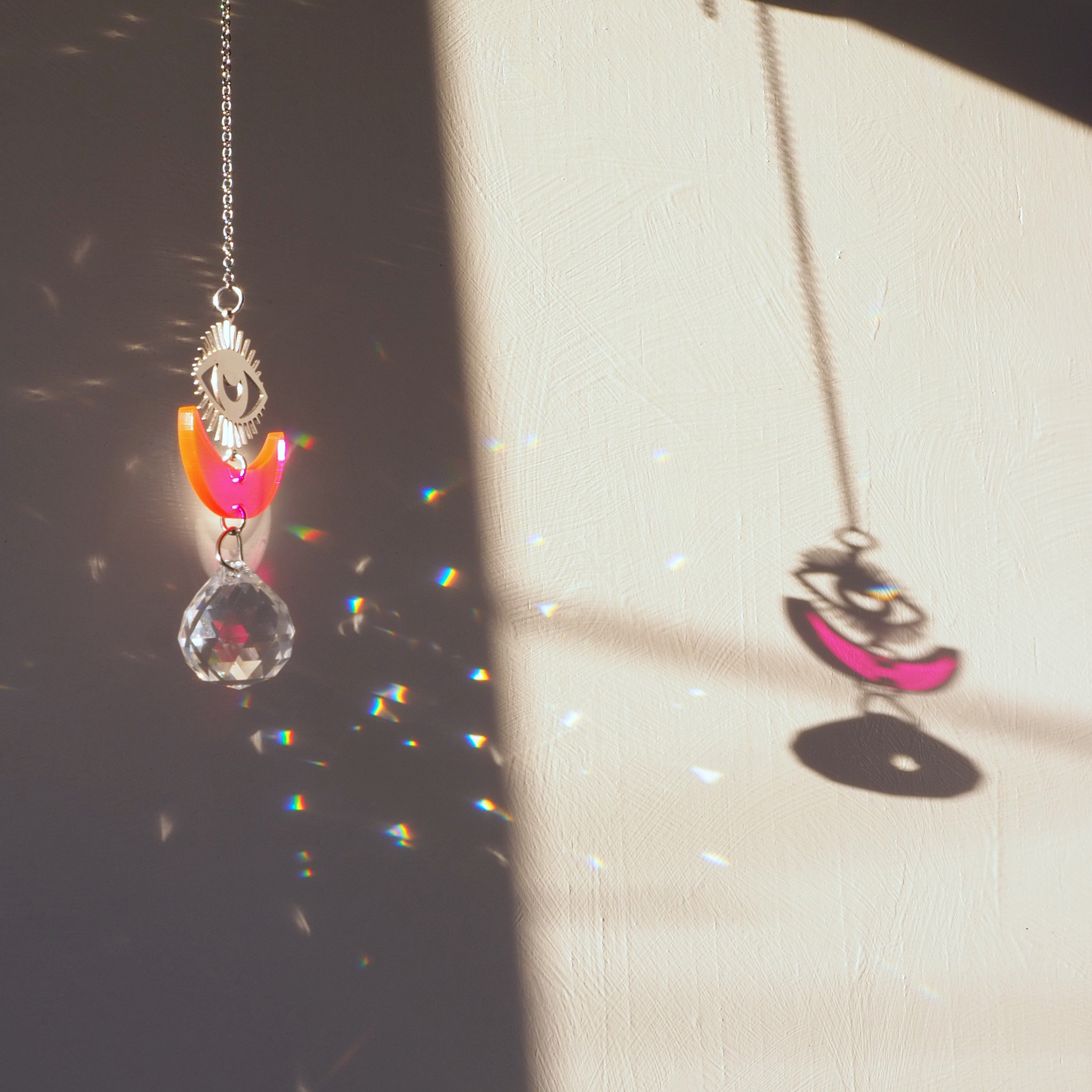 A silver metal eye, neon pink acrylic moon, and faceted prism hanging from a silver chain.
